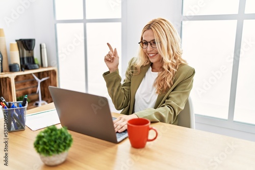 Beautiful blonde woman working at the office with laptop with a big smile on face, pointing with hand and finger to the side looking at the camera.
