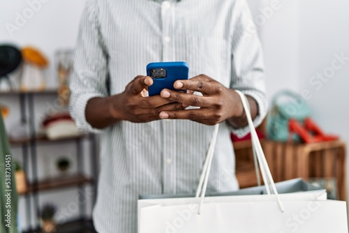 Young african american man using smartphone holding shopping bag at clothing store