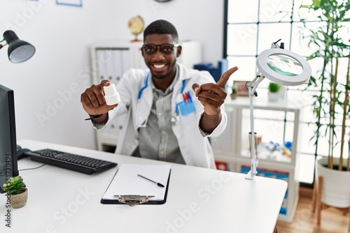 Young african american doctor man wearing doctor uniform holding prescription pills at the clinic smiling happy pointing with hand and finger to the side