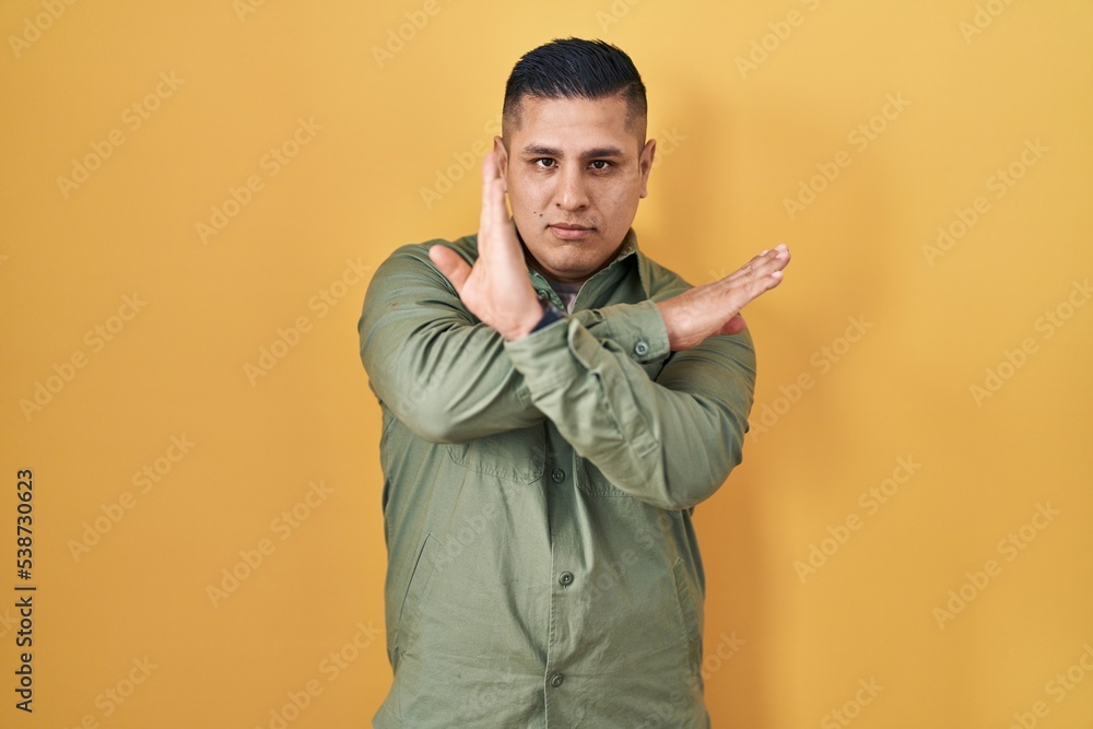 Hispanic young man standing over yellow background rejection expression crossing arms doing negative sign, angry face Hispanic young man standing over yellow background rejection expression crossing arms doing negative sign, angry face