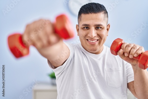 Young latin man smiling confident using dumbbells boxing at home