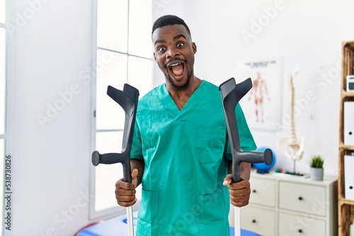 Young african physiotherapist man holding crutches celebrating crazy and amazed for success with open eyes screaming excited.