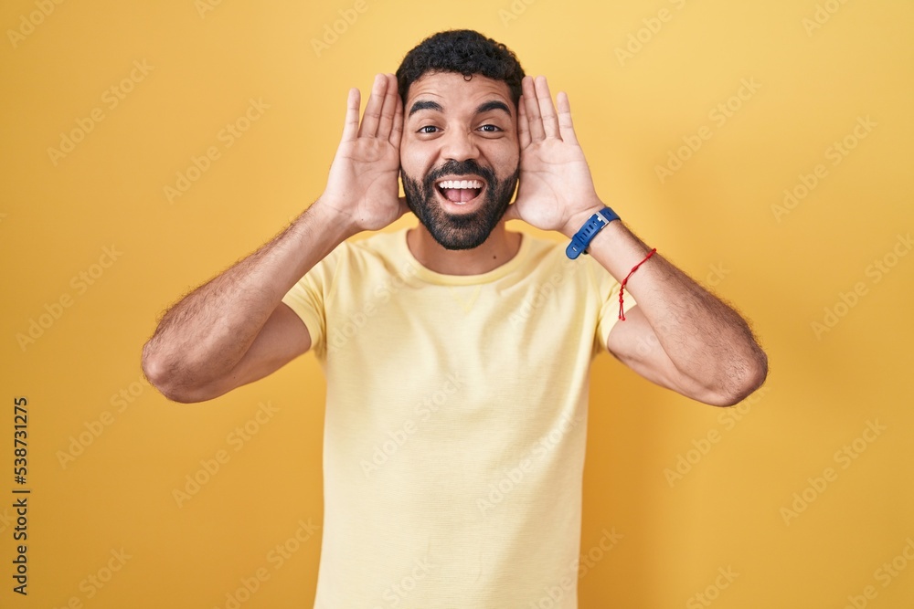 Hispanic man with beard standing over yellow background smiling cheerful playing peek a boo with hands showing face. surprised and exited Hispanic man with beard standing over yellow background smiling cheerful playing peek a boo with hands showing face. surprised and exited