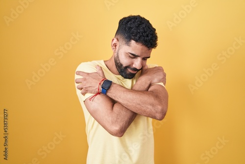 Hispanic man with beard standing over yellow background hugging oneself happy and positive, smiling confident. self love and self care