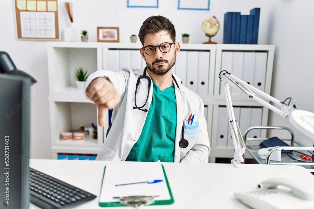 Young man with beard wearing doctor uniform and stethoscope at the clinic looking unhappy and angry showing rejection and negative with thumbs down gesture. bad expression. Young man with beard wearing doctor uniform and stethoscope at the clinic looking unhappy and angry showing rejection and negative with thumbs down gesture. bad expression.