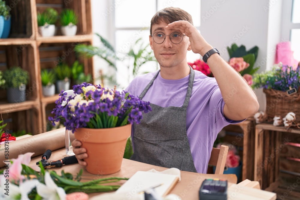 Caucasian blond man working at florist shop very happy and smiling looking far away with hand over head. searching concept.