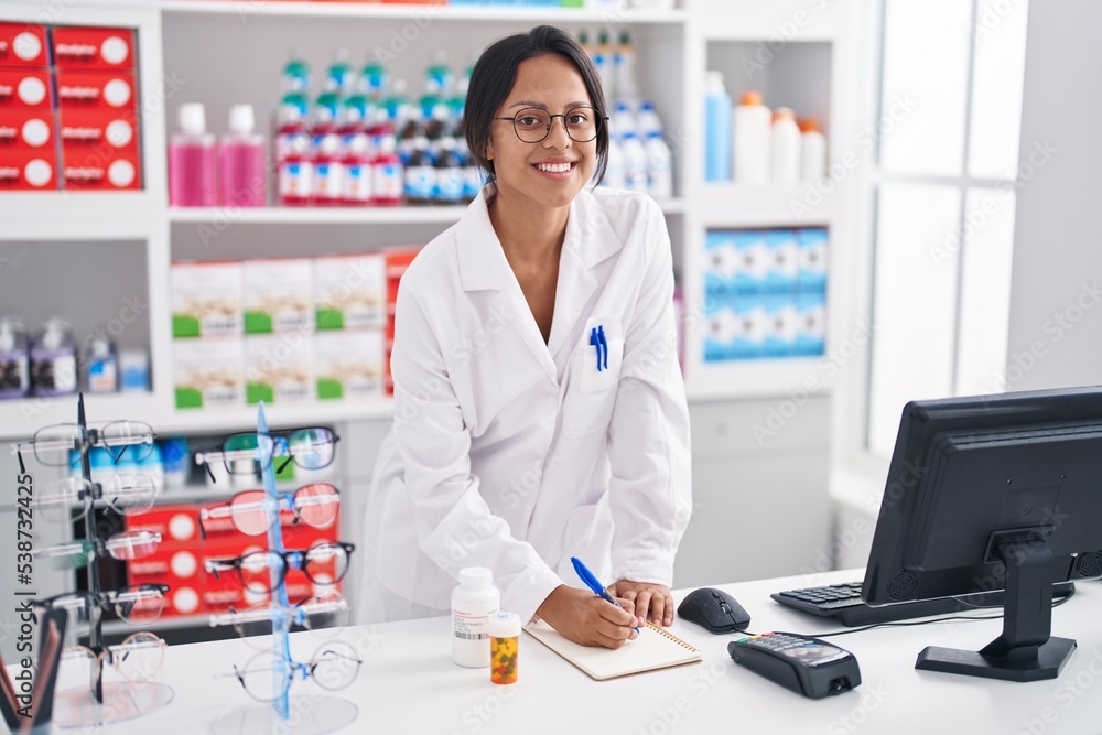 Young hispanic woman pharmacist writing on notebook using computer at pharmacy