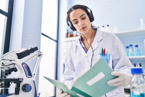 Young hispanic woman wearing scientist uniform listening to music reading book at laboratory