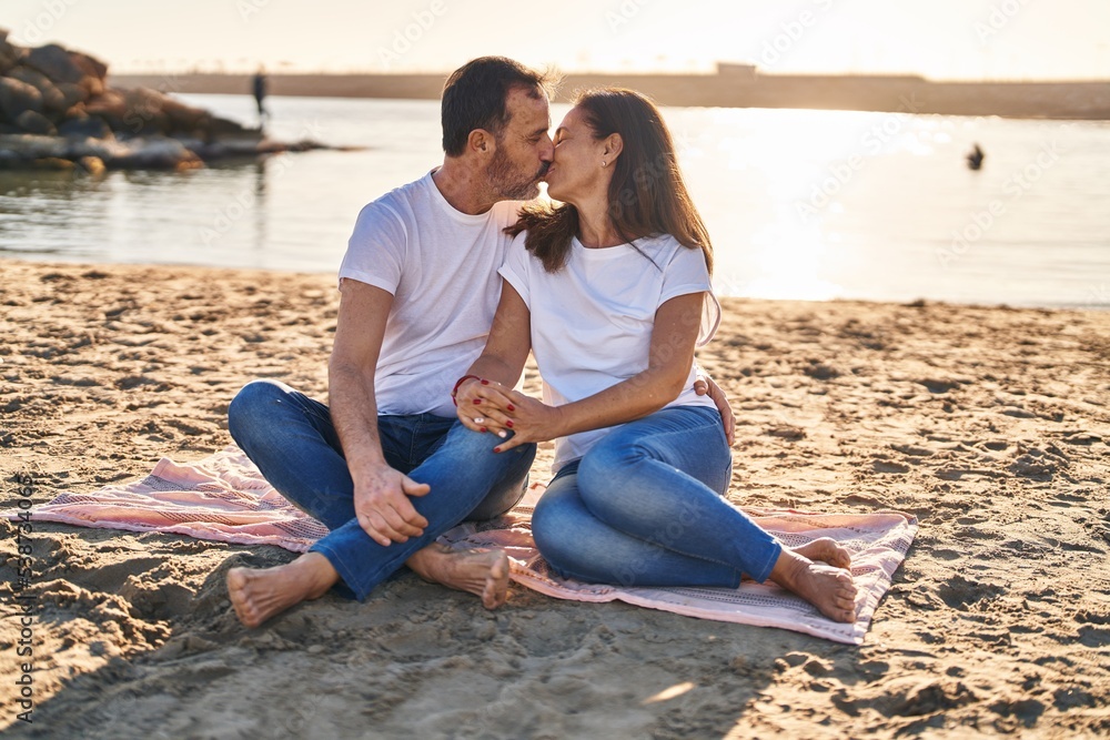 Middle age man and woman couple hugging each other sitting on sand at seaside