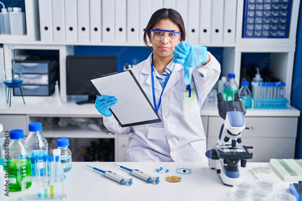 Hispanic young woman working at scientist laboratory looking unhappy and angry showing rejection and negative with thumbs down gesture. bad expression.