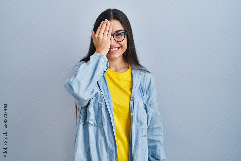 Young hispanic woman standing over blue background covering one eye with hand, confident smile on face and surprise emotion. Young hispanic woman standing over blue background covering one eye with hand, confident smile on face and surprise emotion.