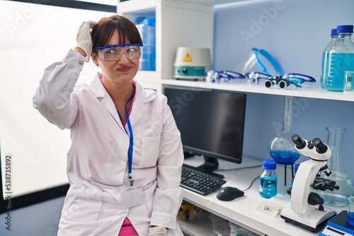 Young brunette woman working at scientist laboratory confuse and wonder about question. uncertain with doubt  thinking with hand on head. pensive concept.