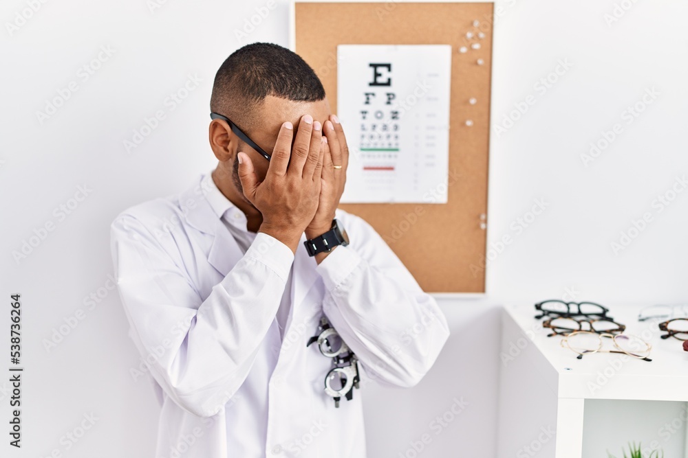 African american optician man standing by eyesight test with sad expression covering face with hands while crying. depression concept.