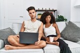 Young interracial couple using laptop at home sitting on the sofa relaxed with serious expression on face. simple and natural looking at the camera.