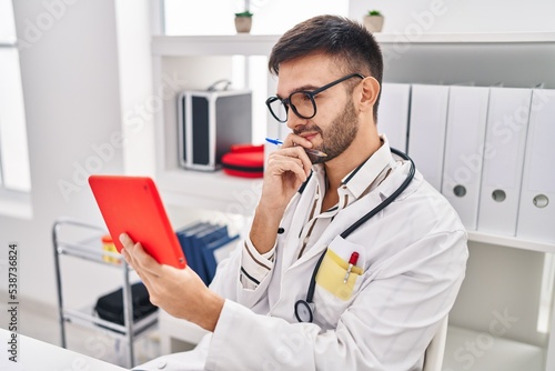Young hispanic man doctor using touchpad sitting on table at clinic