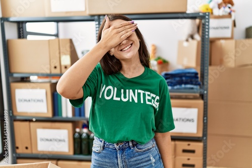 Young brunette woman wearing volunteer t shirt at donations stand smiling and laughing with hand on face covering eyes for surprise. blind concept.