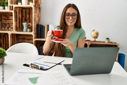 Young hispanic woman using laptop drinking coffee sitting on table at home