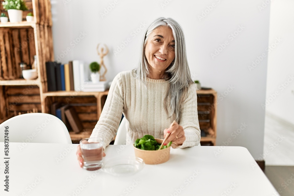 Middle age grey-haired woman eating salad sitting on table at home Middle age grey-haired woman eating salad sitting on table at home