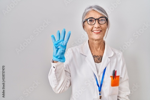 Middle age woman with grey hair wearing scientist robe showing and pointing up with fingers number four while smiling confident and happy.
