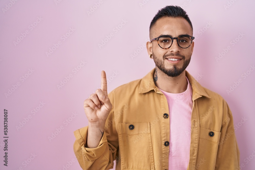 Young hispanic man standing over pink background showing and pointing up with finger number one while smiling confident and happy. Young hispanic man standing over pink background showing and pointing up with finger number one while smiling confident and happy.