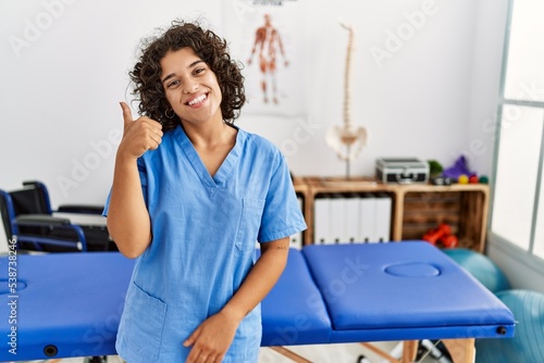 Young hispanic physiotherapist woman working at pain recovery clinic smiling happy and positive  thumb up doing excellent and approval sign