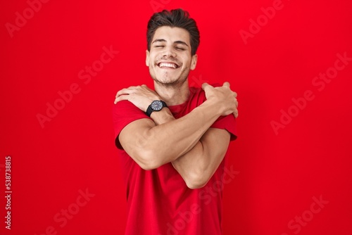 Young hispanic man standing over red background hugging oneself happy and positive, smiling confident. self love and self care