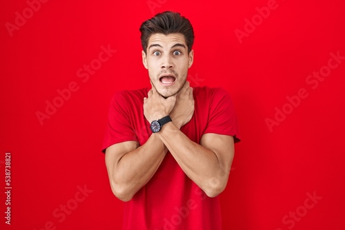 Young hispanic man standing over red background shouting suffocate because painful strangle. health problem. asphyxiate and suicide concept.