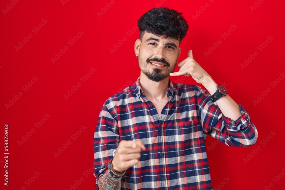 Young hispanic man with beard standing over red background smiling doing talking on the telephone gesture and pointing to you. call me. Young hispanic man with beard standing over red background smiling doing talking on the telephone gesture and pointing to you. call me.