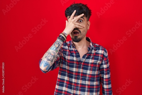 Young hispanic man with beard standing over red background peeking in shock covering face and eyes with hand, looking through fingers with embarrassed expression.