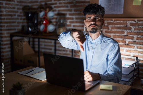 Young hispanic man with beard working at the office at night looking unhappy and angry showing rejection and negative with thumbs down gesture. bad expression.