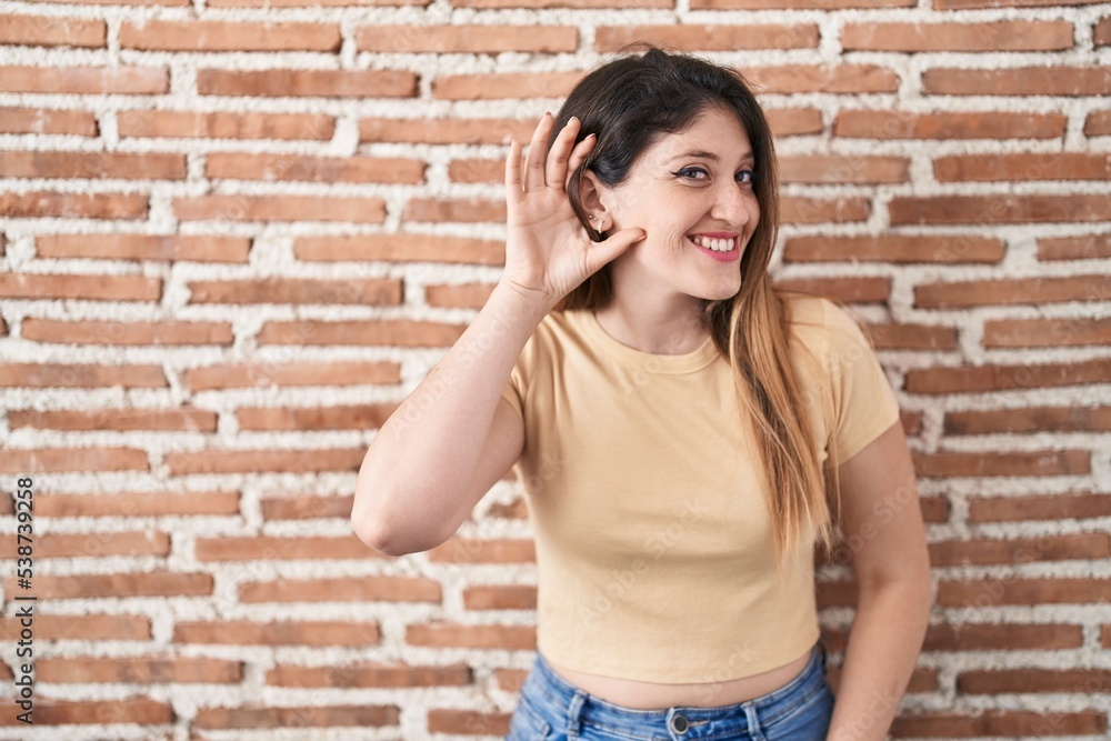 Young brunette woman standing over bricks wall smiling with hand over ear listening an hearing to rumor or gossip. deafness concept. Young brunette woman standing over bricks wall smiling with hand over ear listening an hearing to rumor or gossip. deafness concept.