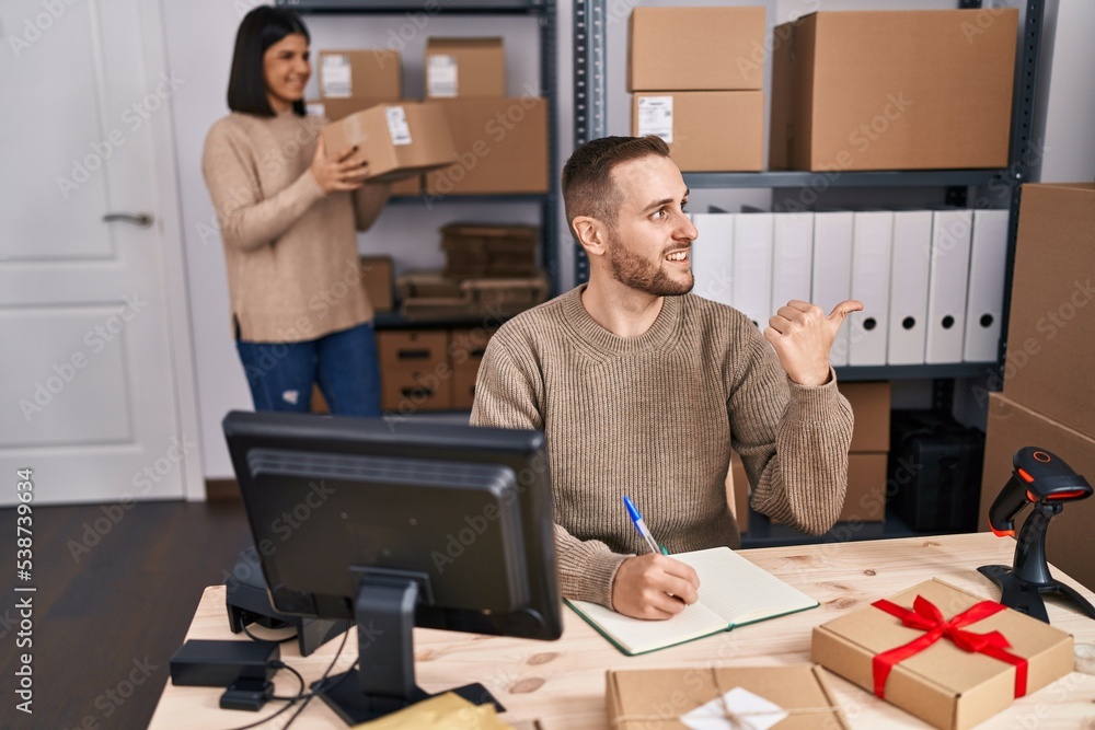 Two young people working at small business ecommerce pointing thumb up to the side smiling happy with open mouth Two young people working at small business ecommerce pointing thumb up to the side smiling happy with open mouth