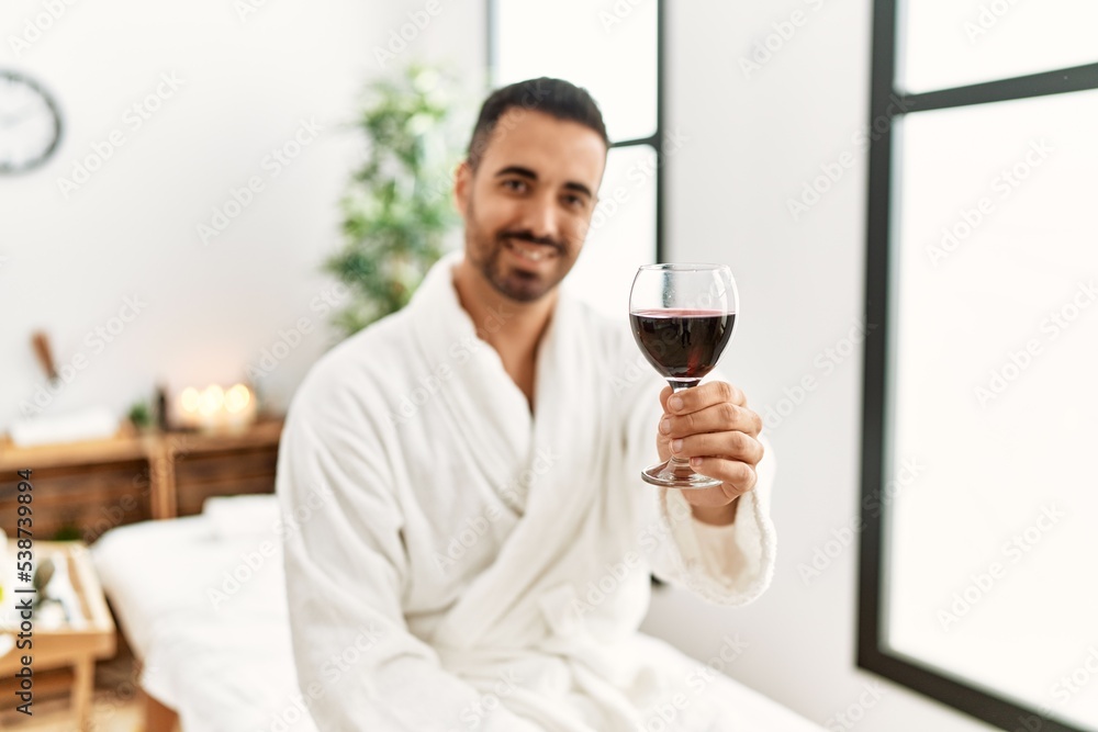 Young hispanic man wearing bathrobe toasting with wine at beauty center Young hispanic man wearing bathrobe toasting with wine at beauty center