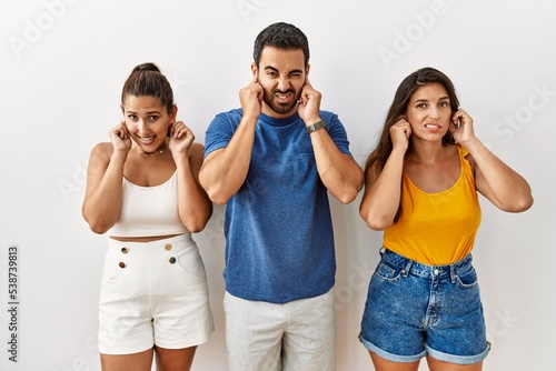 Group of young hispanic people standing over isolated background covering ears with fingers with annoyed expression for the noise of loud music. deaf concept.