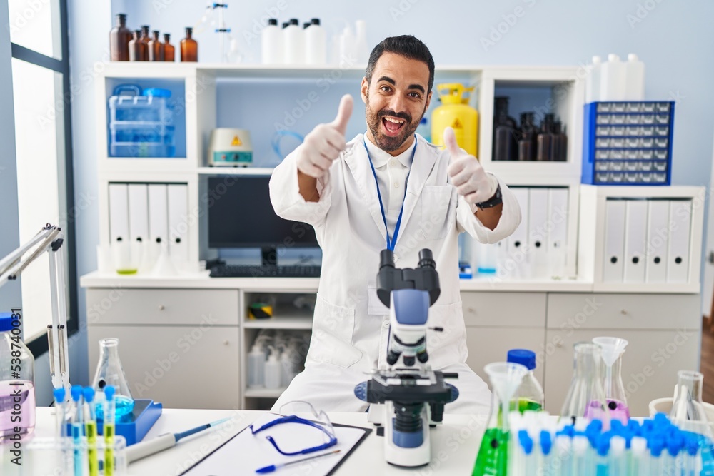 Young hispanic man with beard working at scientist laboratory approving doing positive gesture with hand, thumbs up smiling and happy for success. winner gesture. Young hispanic man with beard working at scientist laboratory approving doing positive gesture with hand, thumbs up smiling and happy for success. winner gesture.