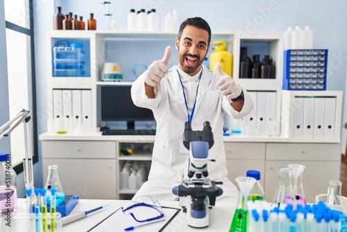 Young hispanic man with beard working at scientist laboratory approving doing positive gesture with hand, thumbs up smiling and happy for success. winner gesture.