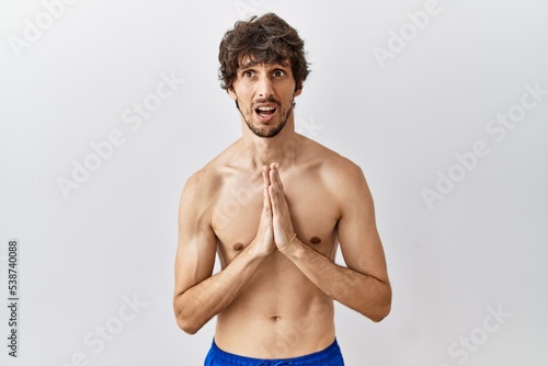 Young hispanic man standing shirtless over isolated, background begging and praying with hands together with hope expression on face very emotional and worried. begging.