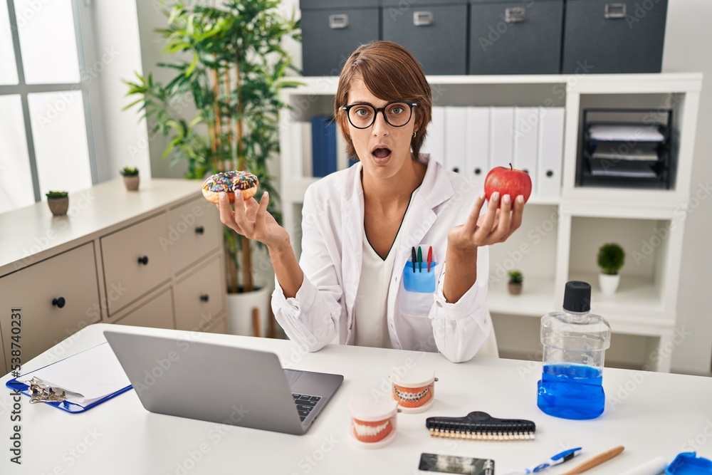 Brunette dentist woman holding apple and chocolate doughnut afraid and shocked with surprise and amazed expression, fear and excited face. Brunette dentist woman holding apple and chocolate doughnut afraid and shocked with surprise and amazed expression, fear and excited face.