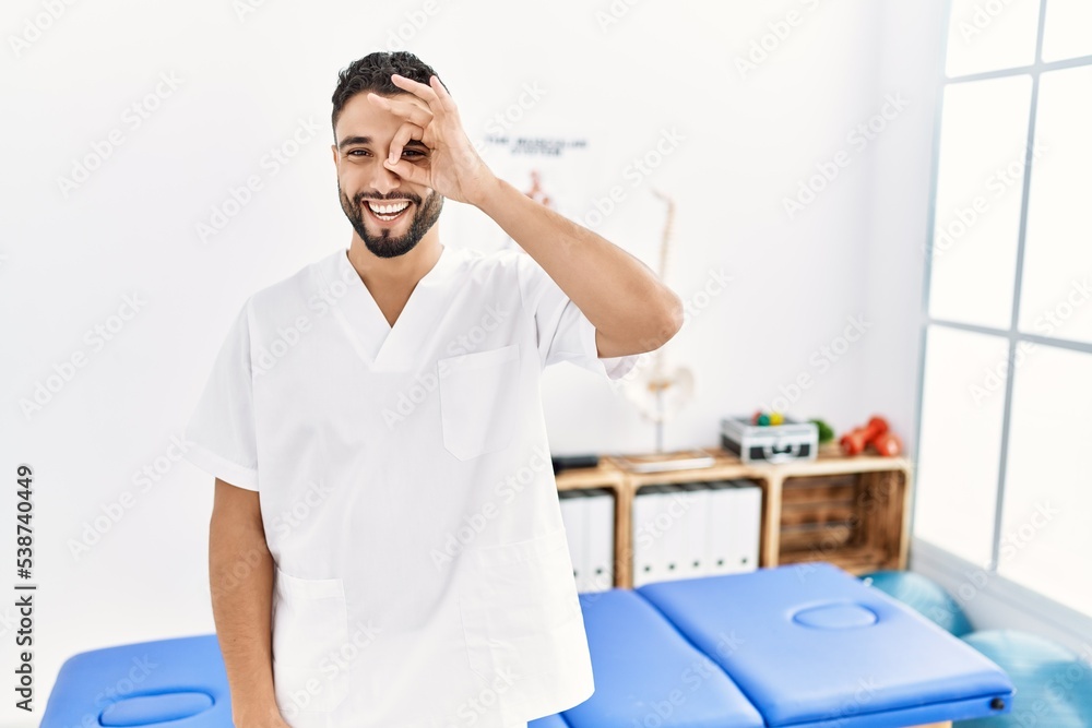 Young handsome man with beard working at pain recovery clinic doing ok gesture with hand smiling, eye looking through fingers with happy face. Young handsome man with beard working at pain recovery clinic doing ok gesture with hand smiling, eye looking through fingers with happy face.