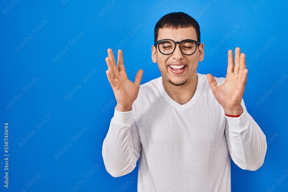 Naklejka premium Young arab man wearing casual white shirt and glasses celebrating mad and crazy for success with arms raised and closed eyes screaming excited. winner concept