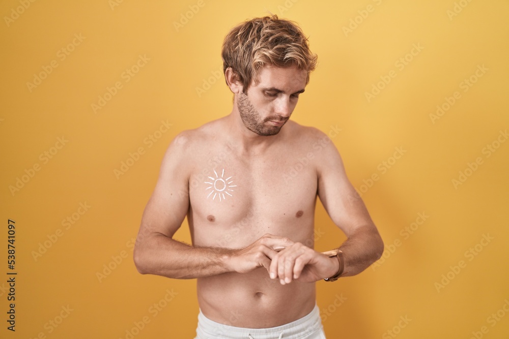 Caucasian man standing shirtless wearing sun screen checking the time on wrist watch, relaxed and confident
