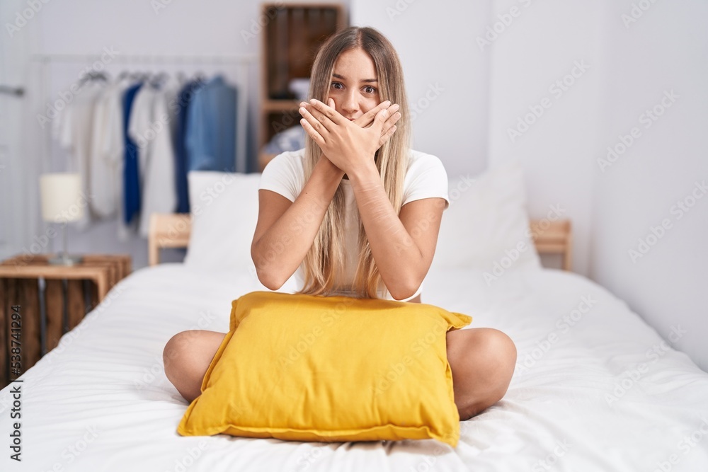 Young blonde woman sitting on the bed with pillow at home shocked covering mouth with hands for mistake. secret concept. Young blonde woman sitting on the bed with pillow at home shocked covering mouth with hands for mistake. secret concept.