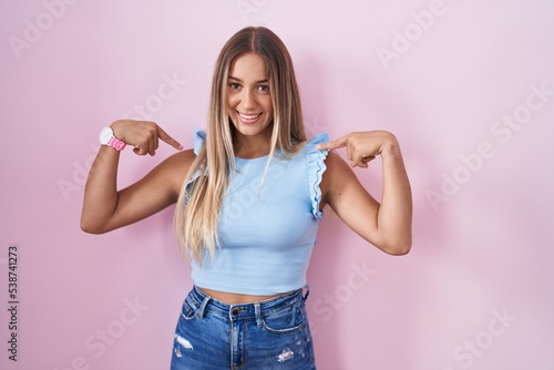 Young blonde woman standing over pink background looking confident with smile on face, pointing oneself with fingers proud and happy.