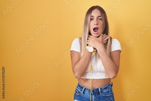 Young blonde woman standing over yellow background wearing headphones shouting suffocate because painful strangle. health problem. asphyxiate and suicide concept.