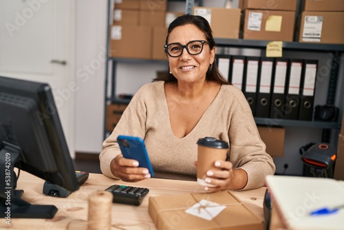 Middle age hispanic woman ecommerce business worker using smartphone drinking coffee at office