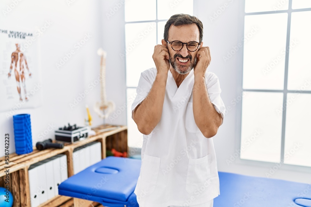 Middle age man with beard working at pain recovery clinic covering ears with fingers with annoyed expression for the noise of loud music. deaf concept. Middle age man with beard working at pain recovery clinic covering ears with fingers with annoyed expression for the noise of loud music. deaf concept.