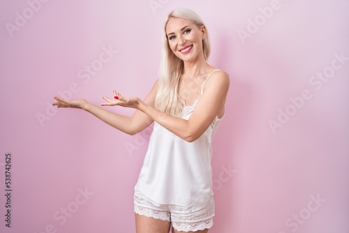 Caucasian woman wearing pajama wearing pink background inviting to enter smiling natural with open hand