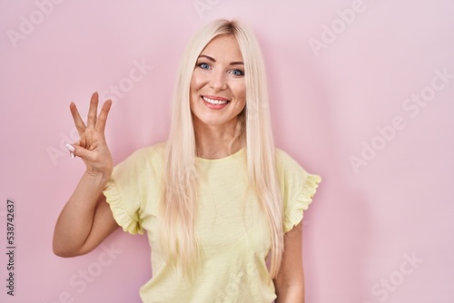 Caucasian woman standing over pink background showing and pointing up with fingers number three while smiling confident and happy.