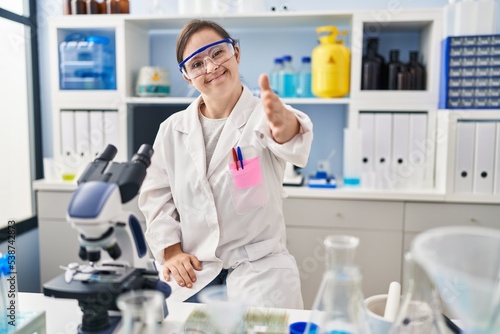 Hispanic girl with down syndrome working at scientist laboratory smiling friendly offering handshake as greeting and welcoming. successful business.