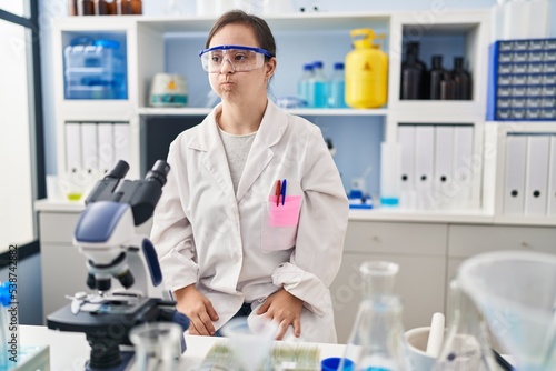 Hispanic girl with down syndrome working at scientist laboratory puffing cheeks with funny face. mouth inflated with air, crazy expression.